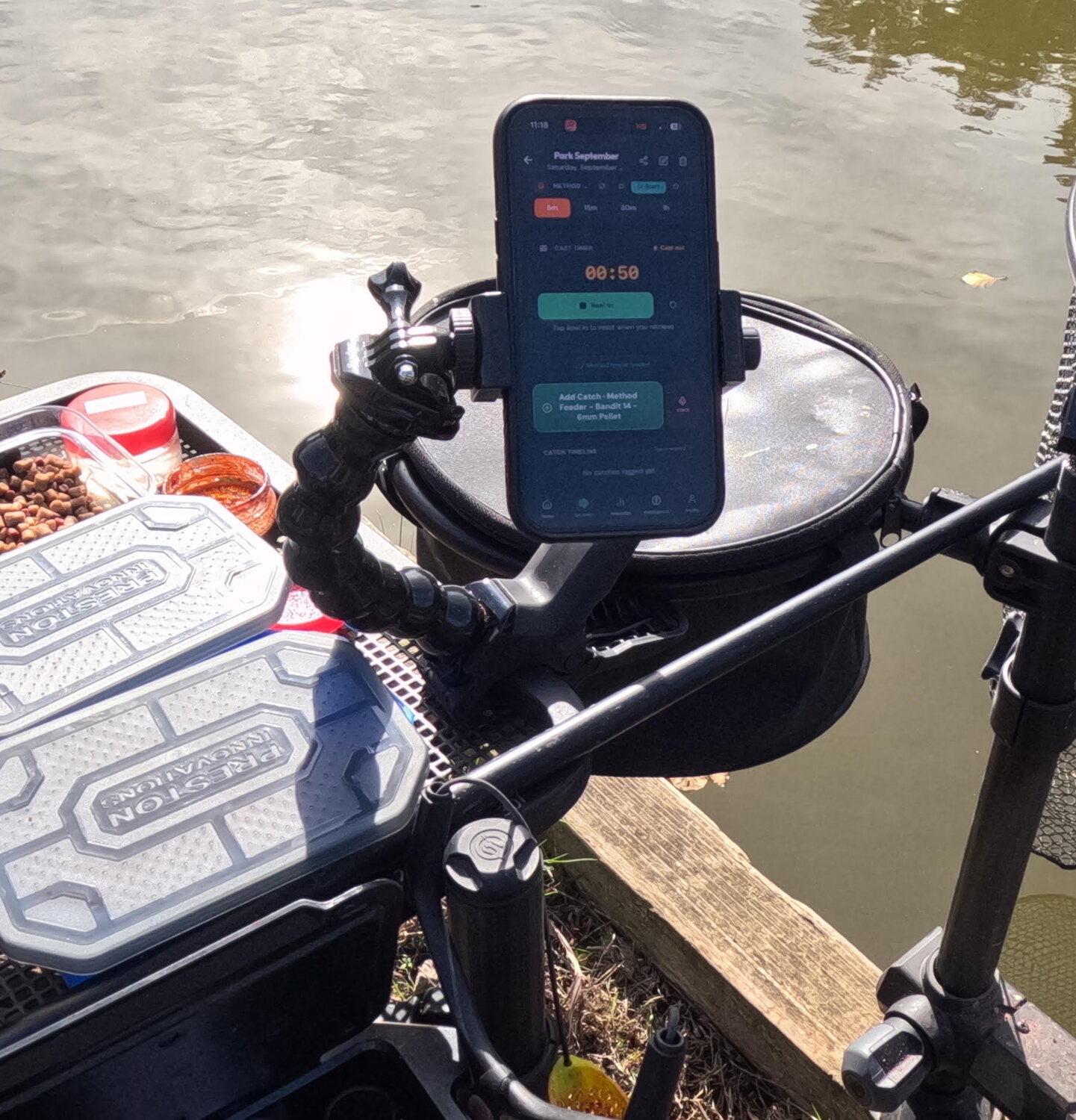 Closer view of a phone in a holder on an articulated flexible arm mounted beside a coarse fishing side tray, with the seat box and tray still available for tackle.