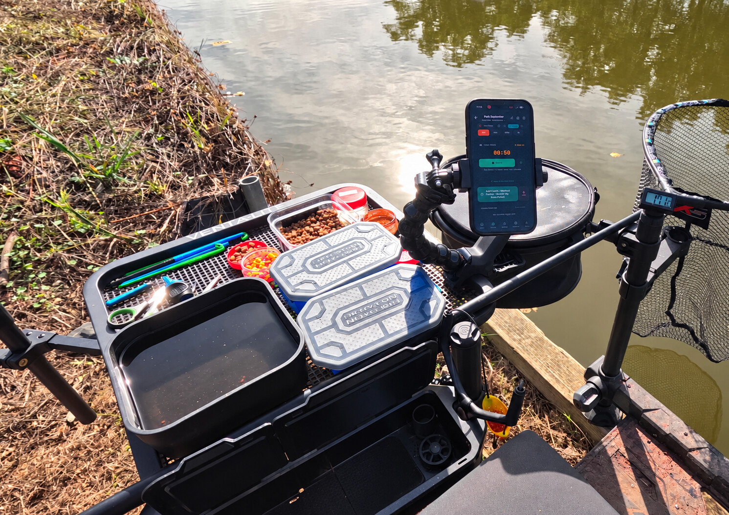 Wide waterside view of a coarse fishing seat box and black mesh side tray set up with bait and tackle, with a phone in portrait orientation on a flexible holder beside the tray and a landing net visible to the right.